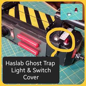 May include: A detailed view of a Ghost Trap prop with a black and yellow striped top, red cylinders, and a switch with a red light. The text "Haslab Ghost Trap Light & Switch Cover" is displayed on a black rectangle.