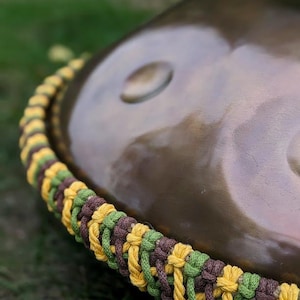 Könnte beinhalten: Ein braunes, metallenes Handpan mit einem gelben, grünen und braunen geflochtenen Seil um den Rand.