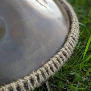 Könnte beinhalten: Nahaufnahme eines braunen Handpans aus Metall mit einem beigefarbenen Seilrand. Das Handpan liegt auf grünem Gras.
