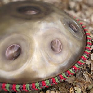 Può includere: Un handpan con finitura metallica e tre fori sonori. L'handpan è decorato con un bordo in corda intrecciata rosso, verde e marrone.