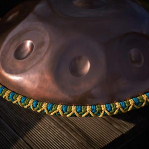 May include: A copper-coloured handpan with a blue and yellow braided rope around the edge.