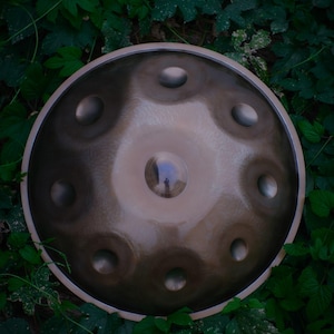 May include: A circular steel handpan drum with a metallic bronze finish, featuring a central dome surrounded by eight circular tone fields arranged in a symmetrical pattern across the convex surface of the percussion instrument.