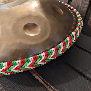May include: A large, round, metallic instrument with a textured surface. The instrument is decorated with a braided rope in red, white, and green.