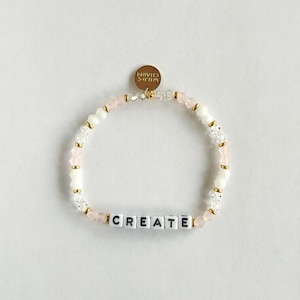 Puede incluir: Una pulsera de cuentas con la palabra "CREATE" escrita con cuentas de letras blancas. La pulsera presenta cuentas rosas, blancas y transparentes, con detalles dorados. Un pequeño dije dorado con las palabras "WILDS CHARM" está adjunto.