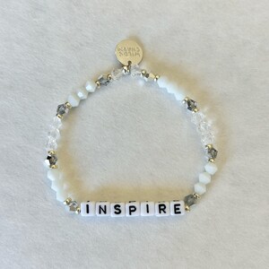 May include: White beaded bracelet with the word "INSPIRE" spelled out in white letter beads. The bracelet also features clear and gray accent beads and a small silver charm that says "WILD'S CHARM".