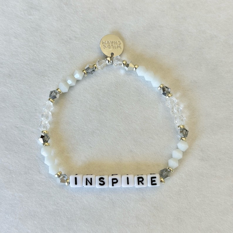 May include: White beaded bracelet with the word "INSPIRE" spelled out in white letter beads. The bracelet also features clear and gray accent beads and a small silver charm that says "WILD'S CHARM".