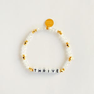 May include: A beaded bracelet with clear and gold-colored beads. The bracelet features white letter beads spelling "THRIVE". A small gold charm with text is attached. The bracelet is on a white background.