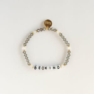 May include: A beaded bracelet with the words "BE KIND" spelled out in white letter beads. The bracelet features gray and clear crystal beads, with a gold charm that says "WILD CHARM".