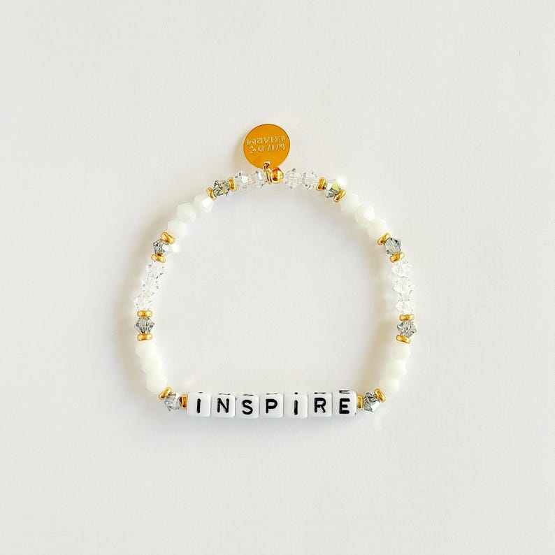 May include: White beaded bracelet with the word "INSPIRE" spelled out in white letter beads. The bracelet has gold accents and a small gold charm. The bracelet is on a white background.