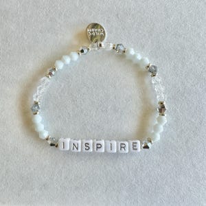 May include: White beaded bracelet with the word "INSPIRE" spelled out in white letter beads. The bracelet also features clear and silver-toned beads, and a small silver charm that reads "WILD CHARM".