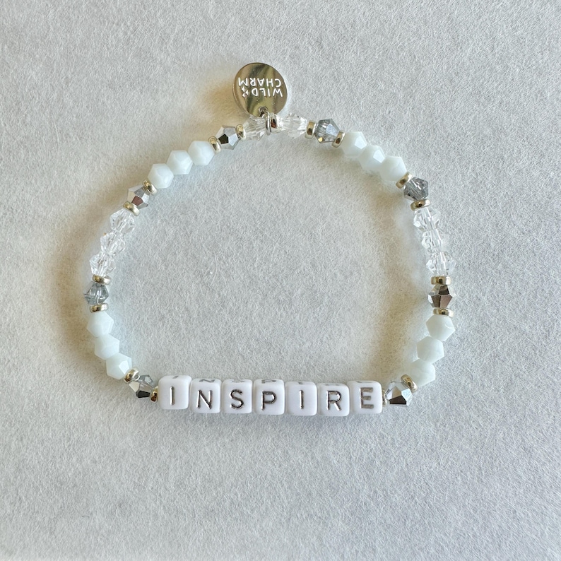 May include: White beaded bracelet with the word "INSPIRE" spelled out in white letter beads. The bracelet also features clear and silver-toned beads, and a small silver charm that reads "WILD CHARM".