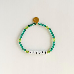 Könnte beinhalten: Ein grünes Perlenarmband mit dem Wort "NATURE" in weißen Buchstabenperlen. Das Armband hat einen goldenen Anhänger mit der Aufschrift "WILD'S CHARM". Die Perlen sind eine Mischung aus facettierten und runden Formen.