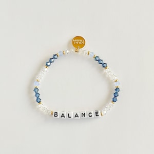 Puede incluir: Una pulsera de cuentas con la palabra "BALANCE" escrita con cuentas de letras blancas. La pulsera presenta cuentas transparentes, azul claro y azul oscuro, con un dije dorado que dice "WILD'S CHARM".