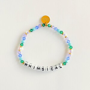 Könnte beinhalten: Ein Armband mit dem Wort "WHIMSICAL" in weißen Buchstabenperlen. Das Armband hat blaue, grüne und goldfarbene Perlen. Ein kleiner goldener Anhänger ist am Armband befestigt.