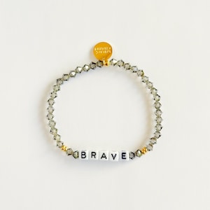 Könnte beinhalten: Ein Armband aus grauen Kristallperlen mit dem Wort "BRAVE" in weißen Buchstabenperlen. Das Armband hat goldfarbene Akzente und einen kleinen goldenen Anhänger. Der Umfang beträgt ca. 18 cm.