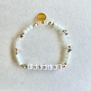 May include: White beaded bracelet with the word "INSPIRE" spelled out in gold letters. The bracelet features clear and silver accent beads, and a gold charm that says "WILD CHARM".