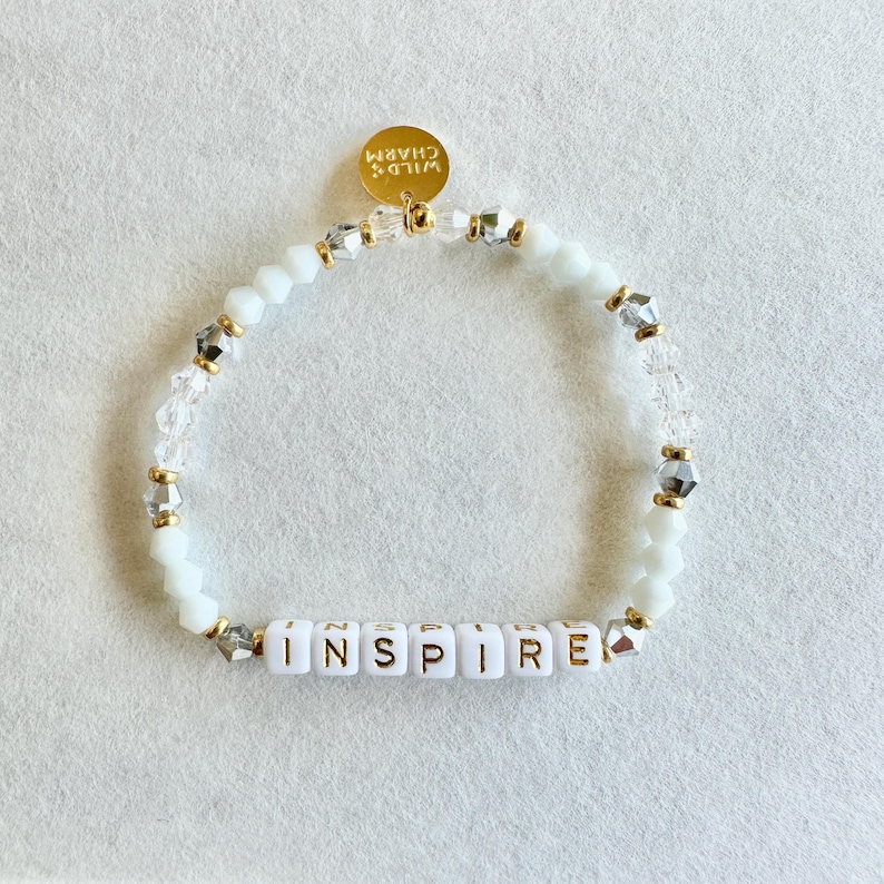 May include: White beaded bracelet with the word "INSPIRE" spelled out in gold letters. The bracelet features clear and silver accent beads, and a gold charm that says "WILD CHARM".