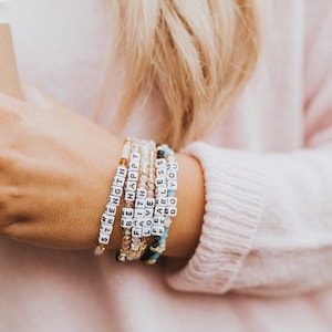 May include: A collection of beaded bracelets with inspirational words. The bracelets feature white letter beads spelling out words like "STRENGTH", "BE HAPPY", and "FEARLESS". The bracelets are in shades of pink, gold, and turquoise.