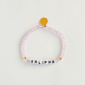 May include: A light pink beaded bracelet with the letters GRL PWR spelled out in white and black letter beads. The bracelet has gold-colored accents and a small gold charm.