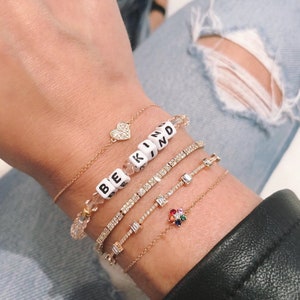 May include: A collection of gold bracelets with various designs. One bracelet spells out "BE KIND" with white letter beads. Other bracelets feature a heart, diamonds, and a colorful flower.