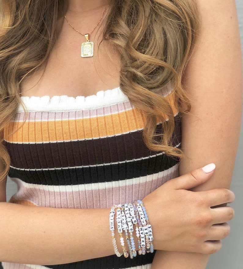 May include: A gold-colored necklace with a square pendant featuring the letter "S" is shown. Several beaded bracelets with white lettered beads spelling out positive words like "Smile", "Dream", and "Hope" are stacked on a wrist. The person is wearing a striped tank top.