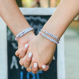 May include: Two beaded bracelets with the words "SOUL SISTER" and "MY PERSON" spelled out in white letter beads. The bracelets are on a person's wrists, with hands clasped together. The bracelets have colorful accent beads.