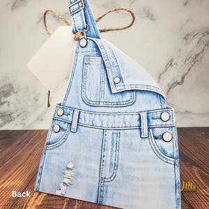 May include: Light blue denim overalls with distressed details, featuring pockets and metal button closures. A blank tag is attached with twine. The overall design is a paper craft or card.