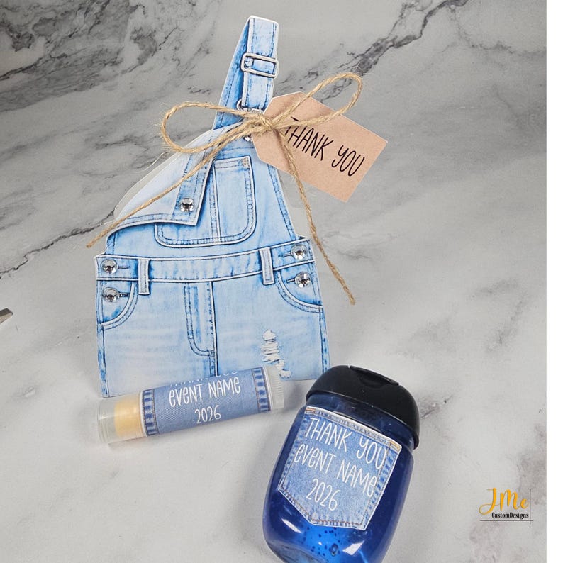 May include: A light blue denim-style gift box shaped like overalls, with a small tag that says "THANK YOU". A lip balm and a blue bottle with the text "THANK YOU EVENT NAME 2026" are also visible.