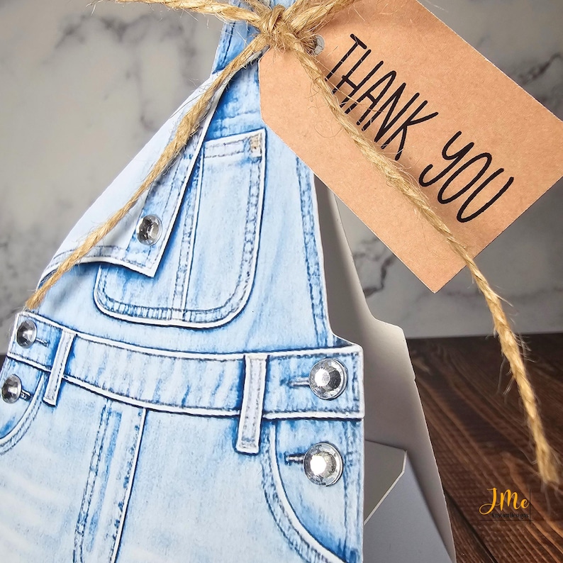 May include: A light blue denim overall-shaped gift box with silver-colored button details. A brown tag tied with twine reads "THANK YOU" in black lettering. The box is partially visible, with a wooden surface in the background.