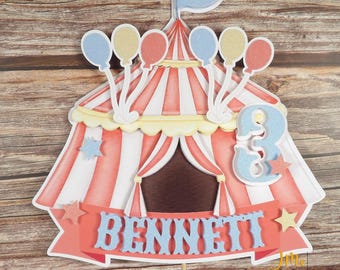 May include: A circus tent cake topper featuring red and white stripes, a blue flag on top, and clusters of glittery blue, yellow, and red balloons. The design includes a blue number 3, a banner with the name BENNETT in blue, and small stars.