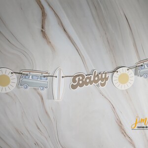 May include: A brown and white paper banner with the word "Baby" in brown letters, a white surfboard, a white van with surfboards on top, and a yellow sun. The banner is strung with twine.
