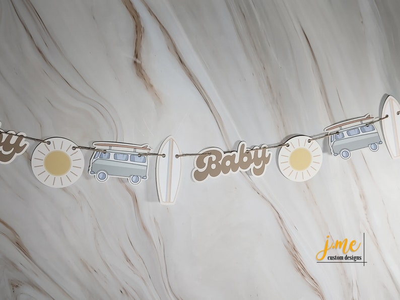 May include: A brown and white paper banner with the word "Baby" in brown letters, a white surfboard, a white van with surfboards on top, and a yellow sun. The banner is strung with twine.