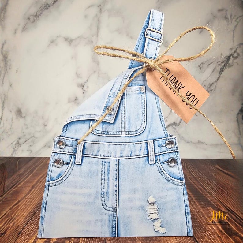 A light blue denim-style gift box shaped like a pair of overalls. The box has distressed details and rhinestone buttons. A brown tag with the words "THANK YOU" is attached with twine.