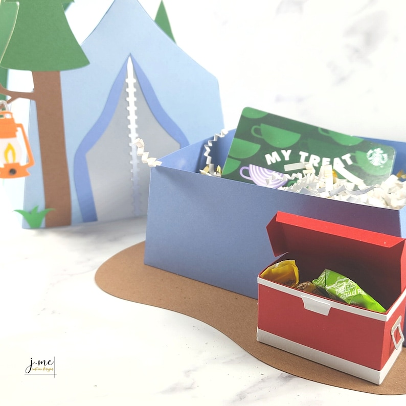 K&ouml;nnte beinhalten: Ein blaues Papier-Campingzelt mit einem braunen Baum und einer gr&uuml;nen Geschenkkarte in einer blauen Box. Ein roter Papierk&uuml;hler mit einem wei&szlig;en Deckel ist ge&ouml;ffnet und enth&auml;lt gr&uuml;ne und gelbe S&uuml;&szlig;igkeiten. Die Szene befindet sich auf einer braunen Papieroberfl&auml;che.