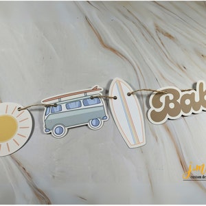 May include: A brown paper banner with the word "Baby" in a retro font. The banner is strung with twine and includes a sun, a vintage van with a surfboard on top, and a surfboard.