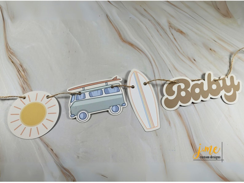 May include: A brown paper banner with the word "Baby" in a retro font. The banner is strung with twine and includes a sun, a vintage van with a surfboard on top, and a surfboard.