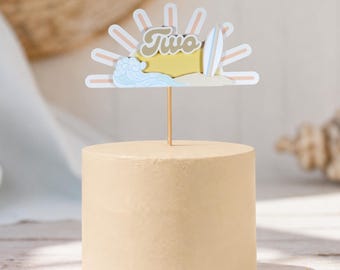 May include: A cylindrical cake with a smooth beige frosting topped with a decorative topper featuring a sunburst design, a blue wave, a surfboard, and the word Two in a retro font, all supported by a thin wooden stick centered on the cake.