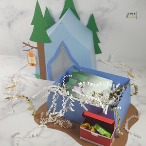 K&ouml;nnte beinhalten: Ein blaues Papierzelt mit einer Geschenkbox im Inneren. Die Geschenkbox ist mit wei&szlig;em Seidenpapier gef&uuml;llt und einer gr&uuml;nen Geschenkkarte mit der Aufschrift "My Treat".