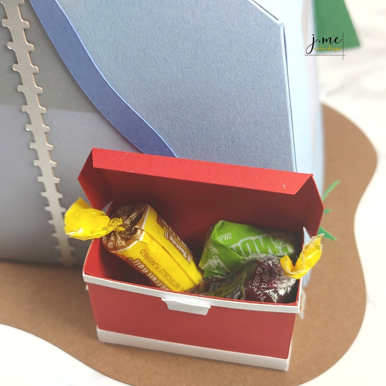 K&ouml;nnte beinhalten: Eine rote Pappbox mit wei&szlig;em Rand, ge&ouml;ffnet, um eine Vielzahl von verpackten S&uuml;&szlig;igkeiten zu enth&uuml;llen. Die S&uuml;&szlig;igkeiten sind in gelbe, gr&uuml;ne und rote Verpackungen verpackt. Die Box steht auf einer braunen Oberfl&auml;che.