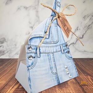 May include: A light blue denim-style gift box shaped like overalls. The box features details like pockets, metal buttons, and distressed denim effects. A brown tag with "THANK YOU" is tied with twine.
