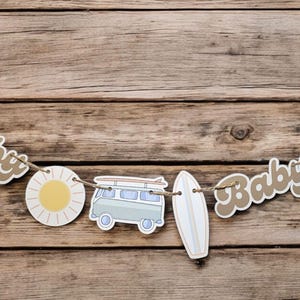 May include: A brown and white baby shower banner with the words "Baby" on it. The banner also features a surfboard, a sun, and a vintage van.