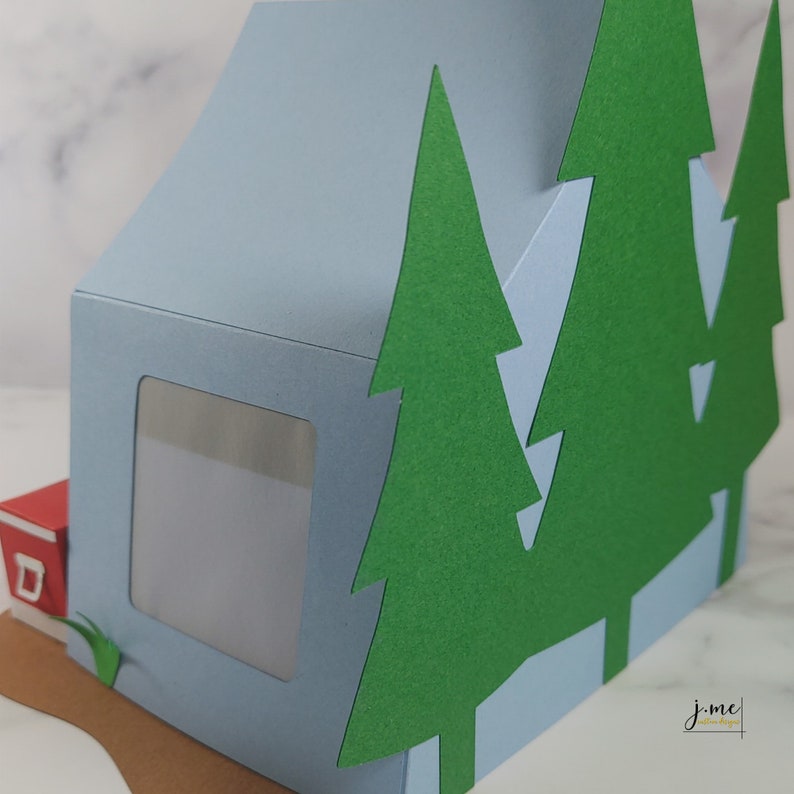 K&ouml;nnte beinhalten: Ein blaues Papierbastelhaus mit einem Fenster und einem roten Briefkasten. Das Haus ist von gr&uuml;nen Papierb&auml;umen umgeben.