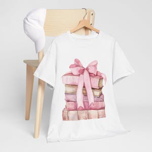 Può includere: T-shirt bianco con un'illustrazione ad acquerello di una pila di libri rosa, legata con un grande fiocco rosa. La maglietta è appesa a una gruccia di legno, con un berretto da baseball bianco su una sedia di legno sullo sfondo.