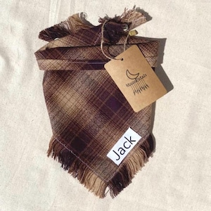 May include: Brown and tan plaid dog bandana with frayed edges. The bandana has a white tag with the name "Jack" on it. The bandana also has a brown tag with the text "Moon Petals" and a crescent moon design.