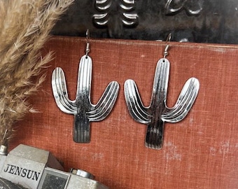 May include: A pair of silver-colored cactus-shaped earrings. The earrings are hanging against a reddish-brown background. The earrings have a detailed design with vertical lines. A vintage camera is visible in the lower left corner.