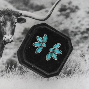 May include: A pair of turquoise flower-shaped earrings in a black velvet box. The earrings are made of silver and have a delicate design.