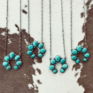 May include: Four silver necklaces with turquoise-colored stone pendants. Each pendant is a horseshoe shape, with the stones set in a silver metal. The necklaces are displayed on a brown and white cowhide-patterned surface.