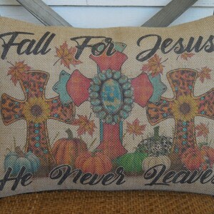 May include: A burlap pillow with a fall-themed design featuring three crosses with turquoise and orange accents. The pillow has the text "Fall For Jesus He Never Leaves" printed on it.