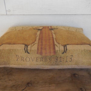 May include: A burlap pillow with a brown and tan design of two sheep standing in front of a tree. The pillow has the text "Proverbs 31:13" printed on the bottom.