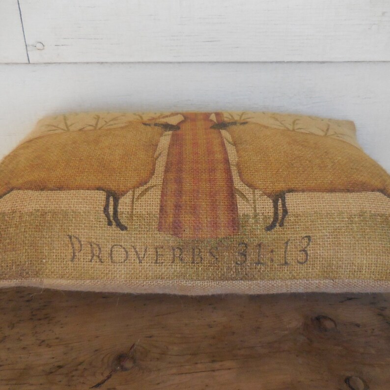 May include: A burlap pillow with a brown and tan design of two sheep standing in front of a tree. The pillow has the text "Proverbs 31:13" printed on the bottom.